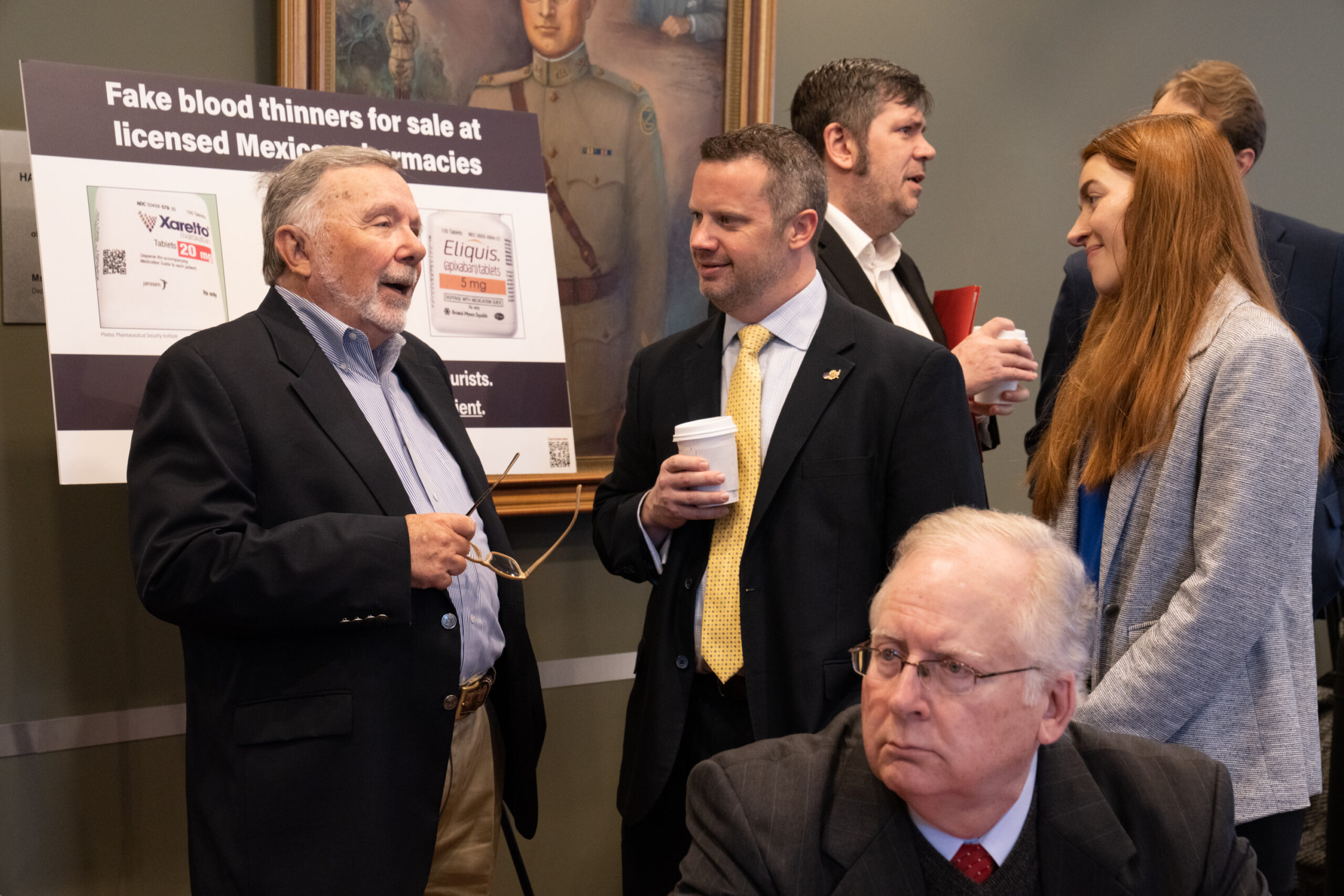 Several people in conversation in front of a military portrait and poster about fake blood thinners in Mexico. A white haired, bespectacled man is sitting in the foreground.