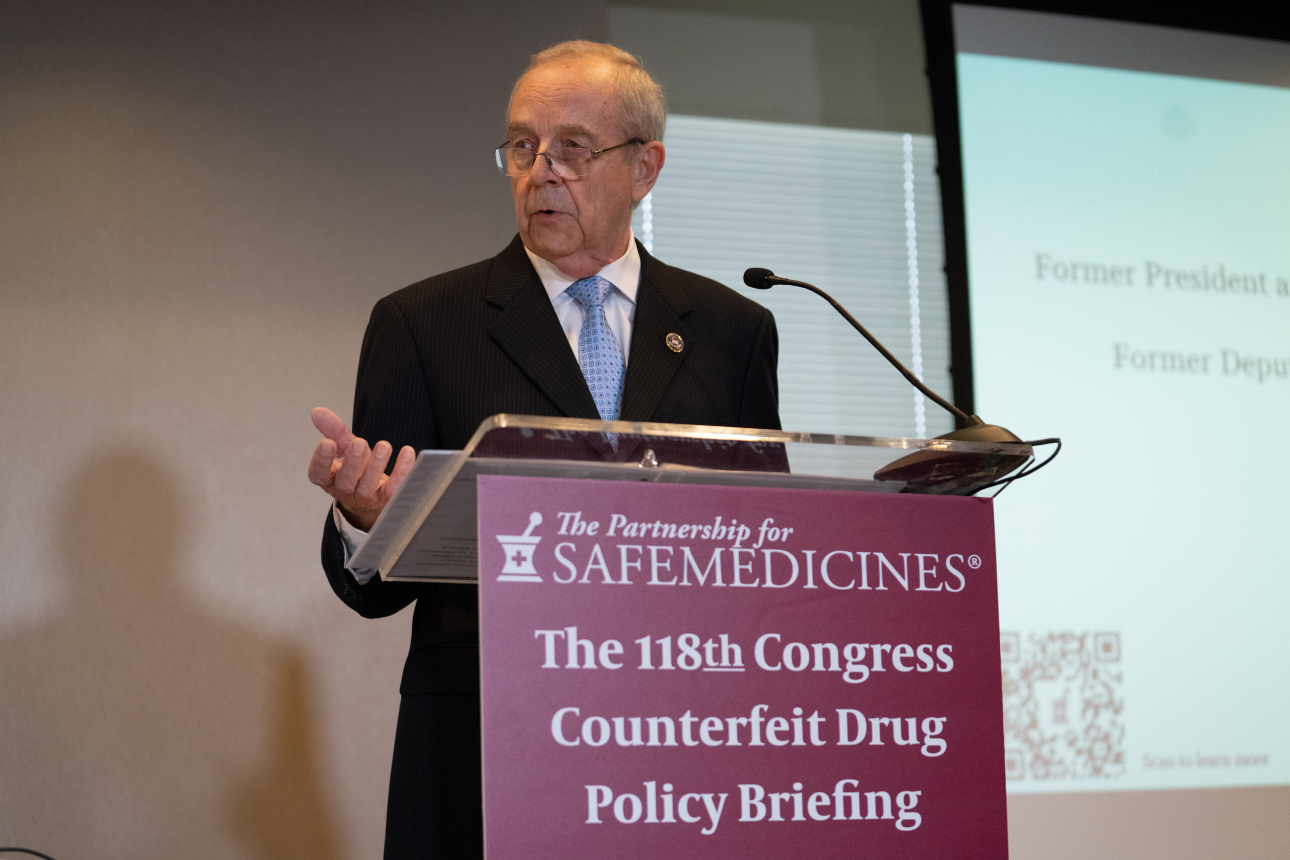 An older man in a dark suit and blue tie at a podium with a sign that reads The Partnership for Safe Medicines, The 118th Congress Counterfeit Drug Policy Briefing.