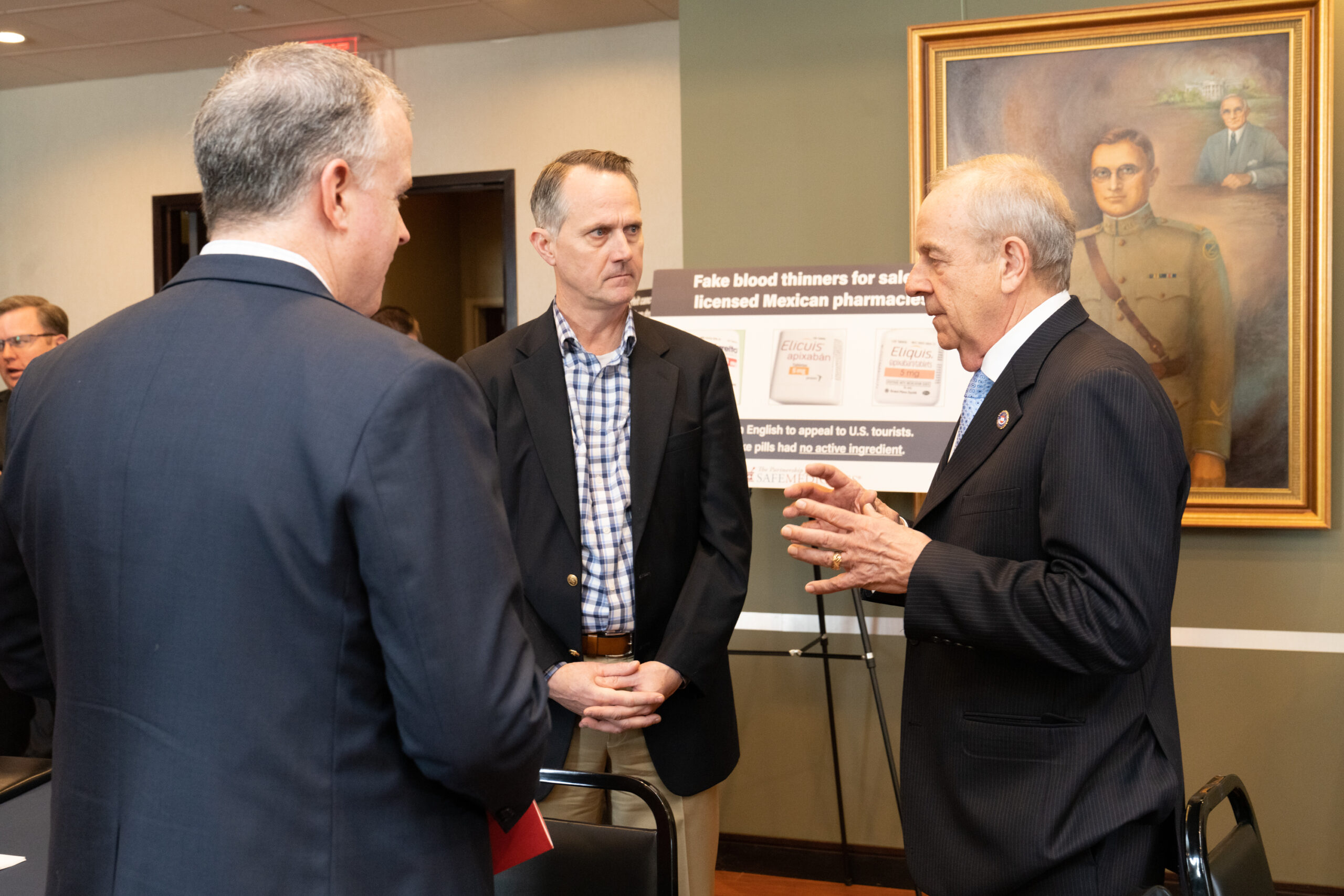 two men in dark suits and a third in a dark sports coat talking in front of a military portrait and a stand up poster about fake blood thinners