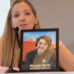A woman in a peach colored sweater talking. In front of her stands a picture of a young teenager labeled Alexander Neville, May 4, 2006 -June 23, 2020