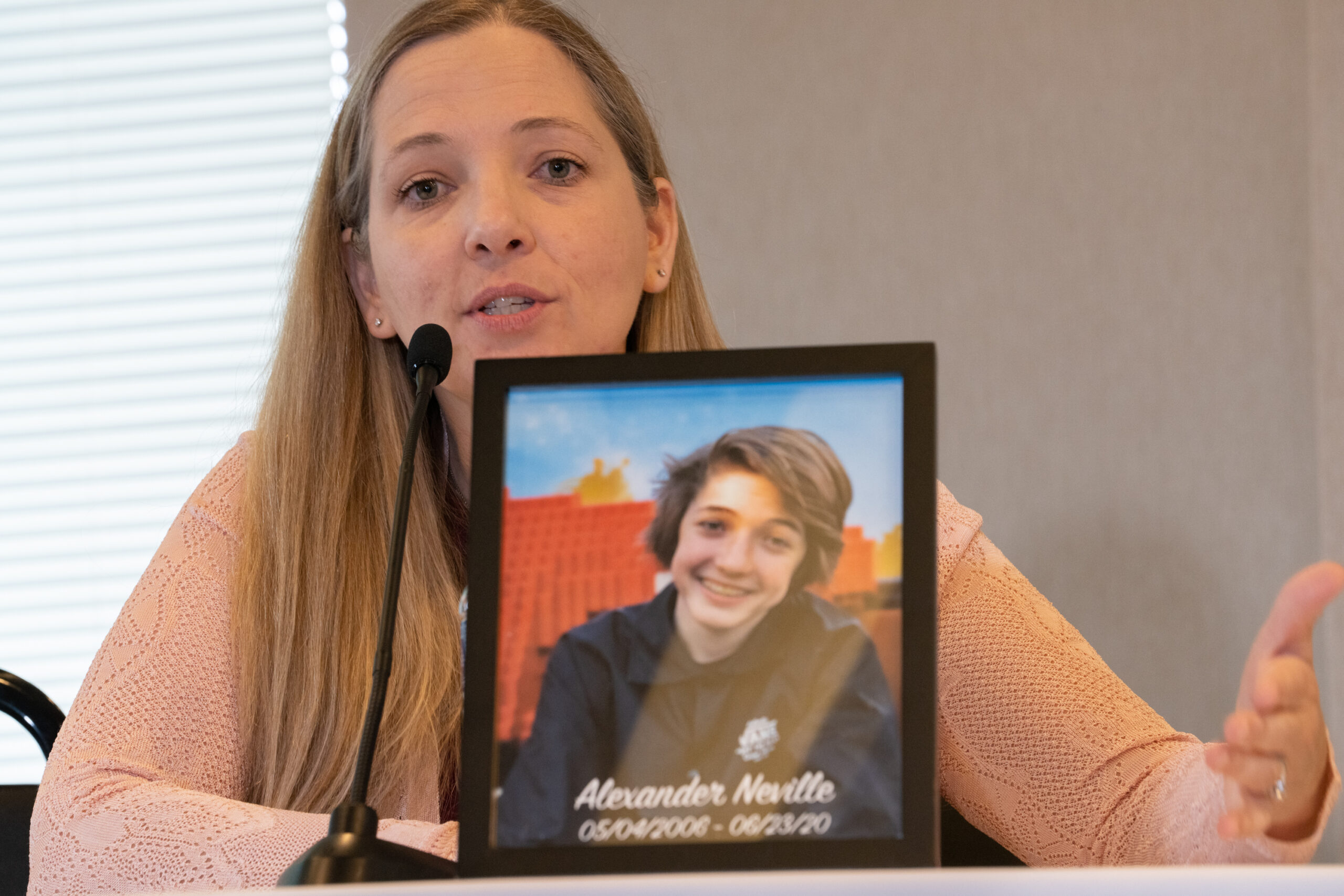 A woman in a peach colored sweater talking. In front of her stands a picture of a young teenager labeled Alexander Neville, May 4, 2006 -June 23, 2020