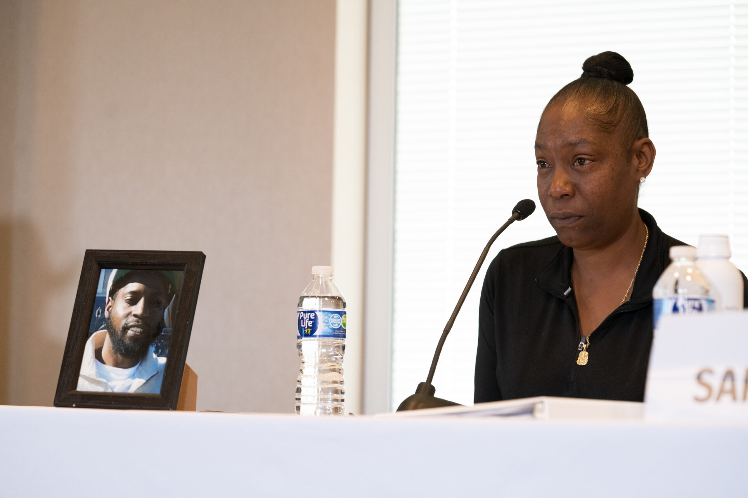 A woman in black with a microphone and a bottle of water in front of her. To her right is a picture of a young, bearded Black man in a white hat and clothes.