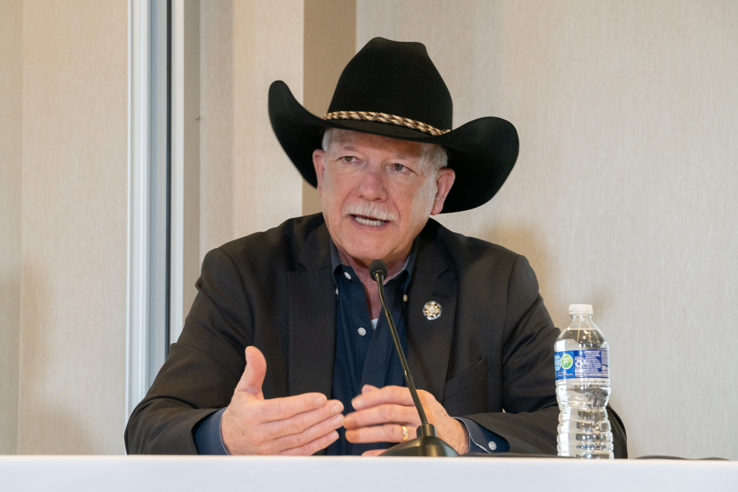 A white haired man in black with a black cowboy hat speaking into a microphone. A bottle of water is to his right.