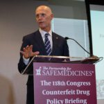 A bald, besuited man with a striped tie at a podium with a sign that reads The Partnership for Safe Medicines, The 118th Congress Counterfeit Drug Policy Briefing. One of his hands is outstretched.