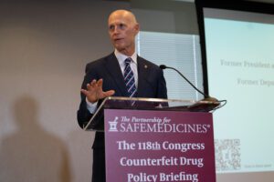 A bald, besuited man with a striped tie at a podium with a sign that reads The Partnership for Safe Medicines, The 118th Congress Counterfeit Drug Policy Briefing. One of his hands is outstretched.
