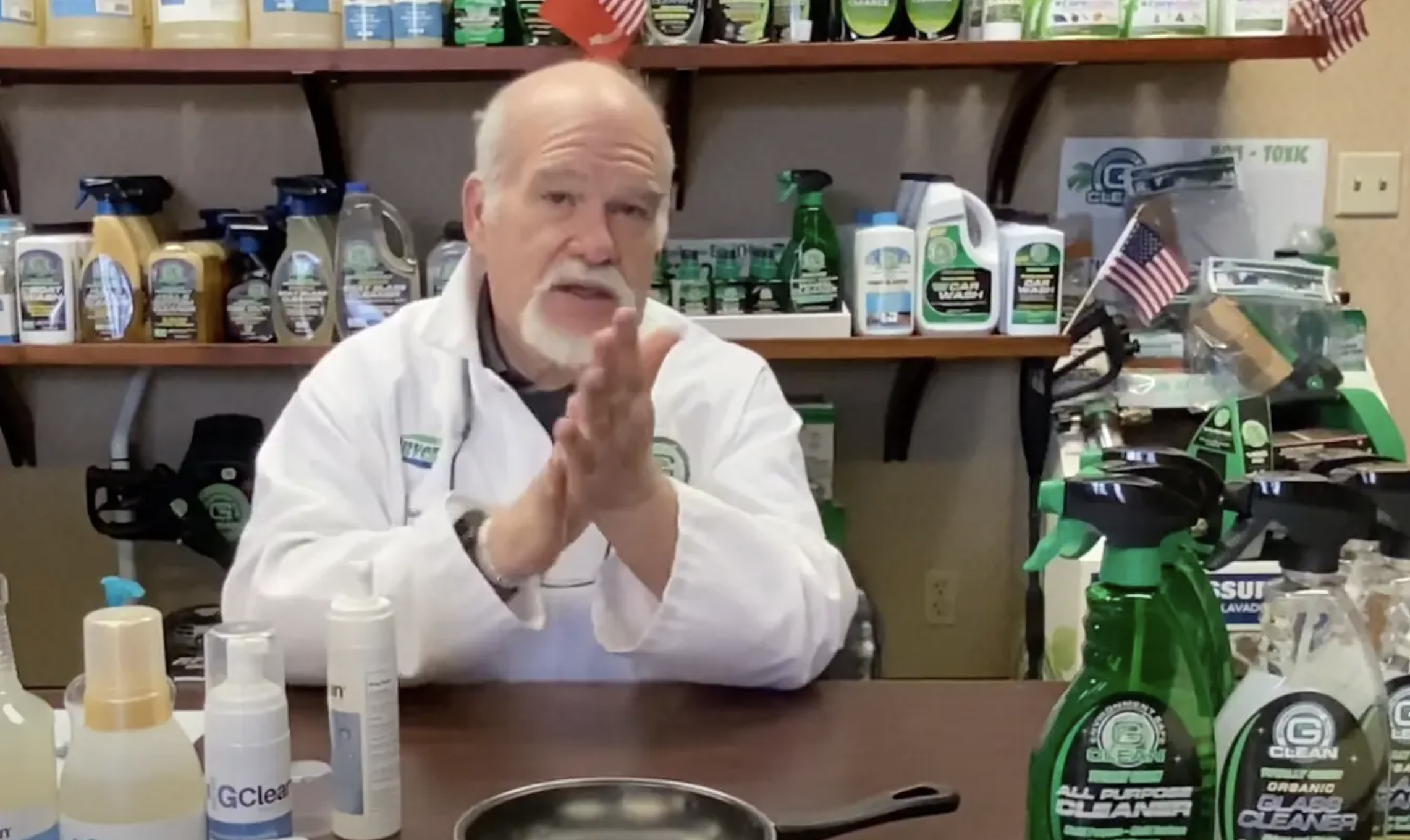 Bearded, white-haired man in labcoat rubbing his hands together in front of many cleaning products.