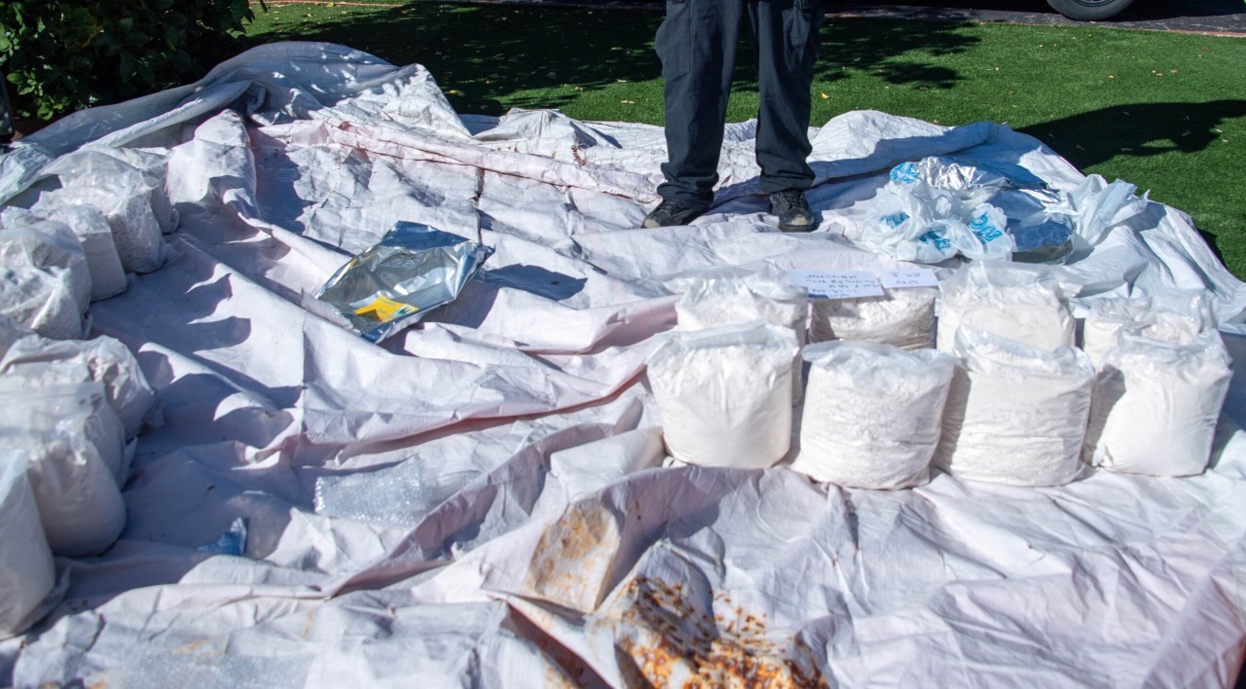 An outdoor picture of clear bags with a white substance in them strewn over a white sheet. someone's show clad feet are visible in the background