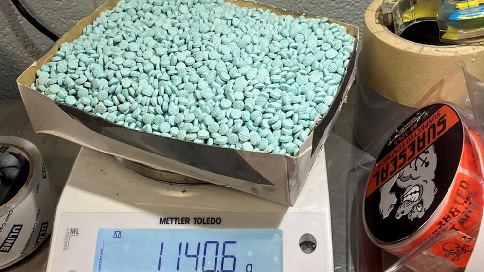 Blue pills in a box top, being weight on a white scale that reads 11406 grams