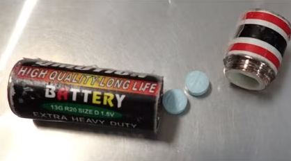 fake AA battery with a screw off top, and two blue pills on a metal surface