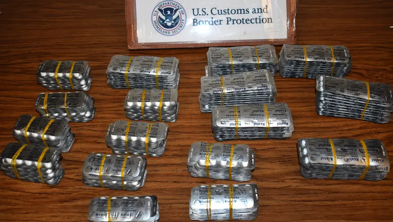 Stacks of foil blister packs containing pills on a wooden table. A placard in the background reads U.S. Customs and Border Protection