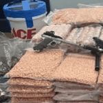 A seizure photo with guns stacked on bags of orange pills. A plastic ACE hardware branded bucket is in the left backgound