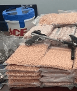 A seizure photo with guns stacked on bags of orange pills. A plastic ACE hardware branded bucket is in the left backgound